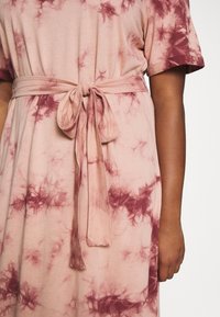 Vaaleanpunainen mekko, jossa on marooni sidontakuvio. Lyhyet hihat, pehmeä kangas ja kankainen vyö sidottuna vyötärölle. Minimalistinen muotoilu.