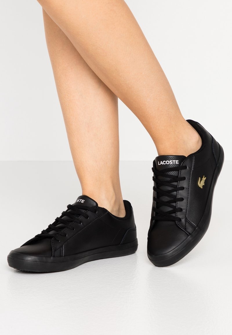 Baskets noires à lacets avec une texture en cuir lisse, arborant un logo Lacoste en or et un design minimaliste.