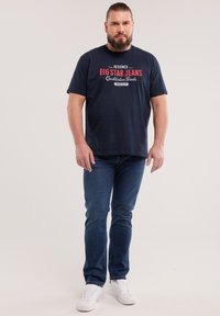 Habemega mees, kes kannab laheda tekstiga tumesinist t-särki, siniseid teksapükse ja valgeid vabaajajalatsid, seisab tavalise heleda tausta ees.