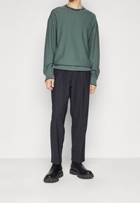 Personne portant un pull vert à manches longues avec le logo Hugo sur le col, un pantalon noir et des bottes noires épaisses sur un fond uni.