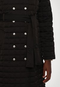 Melna quiltota mēteli ar sudraba pogām, jostu viduklī un strukturētu šuvumu. Mīksta tekstūra ar nedaudz spīdīgu apdari.