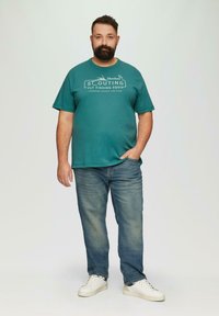 Tealowa bawełniana koszulka z białym nadrukiem, zestawiona z jasnoniebieskimi dżinsami i białymi trampkami. Prosty, luźny krój.