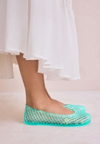 Permatomos tirkizinės balerinos su tinklinio rašto dizainu ir vientisa padais, derančios prie lengvos baltos suknelės. Minkšta tekstūra ir apvalios kojų vietos forma.