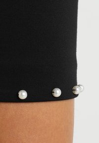 Textile noir avec un gros plan d'un ourlet de manche comportant trois perles blanches décoratives attachées par des coutures noires.