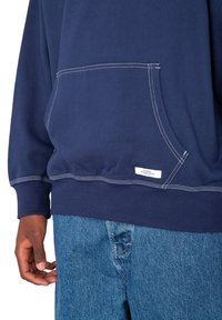 Navy cotton sweatshirt with a kangaroo pocket, white stitching accents, and a small label at the hem. Paired with light blue denim jeans.