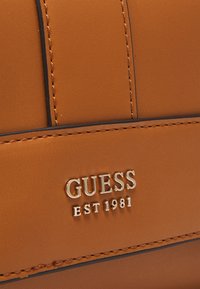 Bruin synthetisch materiaal met gestikte accenten en een goudkleurig "GUESS EST 1981" logo dat prominent wordt weergegeven. Glad textuur en minimalistisch ontwerp.