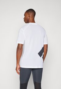 Hvit atletisk T-skjorte med mesh-tekstur, med en svart geometrisk detalj på baksiden. Kortermet og rund hals.