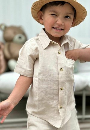 Beige linned skjorte med korte ærmer og en brystlomme, prydet med lyse knapper. En stråhat fuldender outfittet.