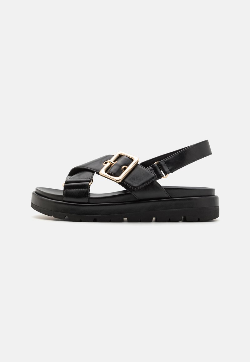 Black sandals with a chunky sole, cross-strap design, and a large gold buckle accent, made from smooth leather with a textured finish.