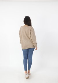 Beige gebreide trui met een losse pasvorm, brede mouwen en geribde onderrand, gecombineerd met blauwe skinny jeans en transparante hakken sandalen.