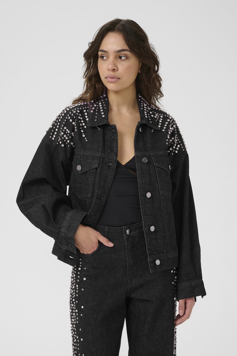 Mujer vestida con chaqueta de mezclilla negra y pantalones decorados con tachuelas plateadas, posando con una mano en el bolsillo, mirando hacia un lado contra un fondo gris.