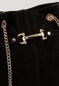 Détail d'un sac à main en daim noir avec une bandoulière chaîne dorée et une fermeture décorative à barre dorée.