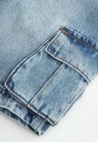 Jasnoniebieska tkanina denimowa prezentująca złożoną kieszeń z kontrastowym brązowym przeszyciem. Tekstura jest gładka z niewielkimi wariacjami w kolorze.