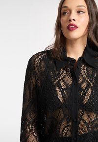 Czarna koronkowa bluzka z misternymi kwiatowymi wzorami, zapinana na guziki, z kontrastującym kołnierzykiem. Przezroczysta tkanina z fakturalnymi zdobieniami.