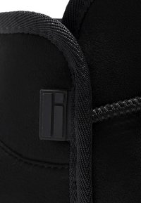 Botín negro con una superficie texturizada, que presenta un acento liso cerca de los cordones y un detalle de logo en forma rectangular en el lateral.