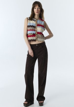 Joven mujer de pie con la mano en la cadera, vestida con un chaleco de punto sin mangas multicolor con zigzag y pantalones anchos oscuros con pequeños puntos.