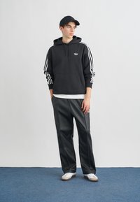 Čierna mikina s kapucňou a bielym logom Adidas a pásikmi na rukávoch, v kombinácii s elegantnými čiernymi nohavicami, bielymi teniskami a zodpovedajúcou čiapkou.