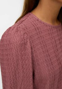 Gros plan sur une personne portant un haut mauve texturé à manches longues avec des détails de couture verticaux et un col rond.