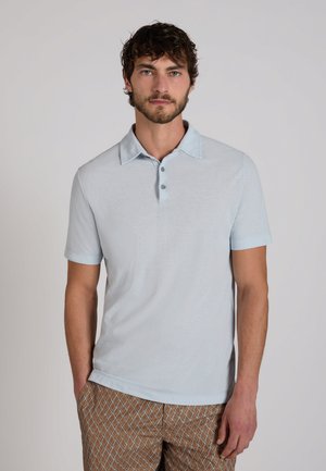 Homme aux cheveux courts et bouclés portant un polo à manches courtes bleu clair et un pantalon marron à motif, debout devant un fond uni.