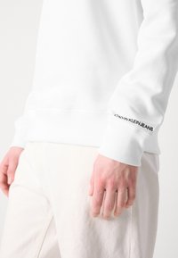 Main droite reposant à côté, portant une chemise blanche à manches longues avec le texte "Calvin Klein Jeans" sur le poignet, associée à un pantalon de couleur claire.