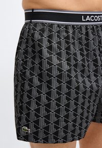Boxer noir avec motif géométrique, logo Lacoste sur la cuisse gauche et ceinture élastique de marque, porté par une personne.