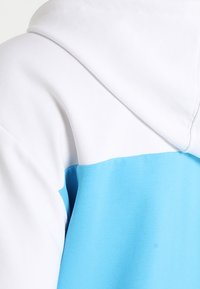 Felpa con cappuccio caratterizzata da una parte superiore bianca e una parte inferiore azzurra uniforme, realizzata in tessuto morbido con una superficie liscia.