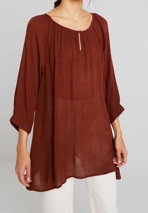 Frau trägt eine lockere, langärmelige, rostfarbene Bluse mit kleinem Schlitz und Knopfdetail am Ausschnitt, kombiniert mit weißen Hosen.