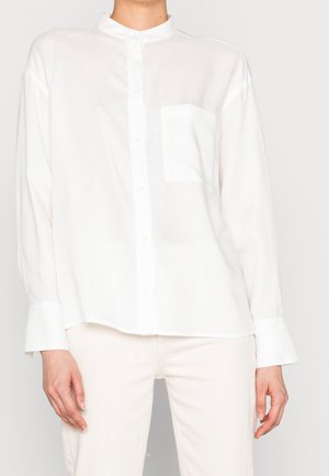 Persoon draagt een loszittend wit overhemd met knopen en een borstzak, gecombineerd met een witte broek, staand tegen een effen lichte achtergrond.