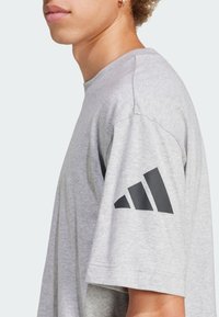 Grå bomulls-t-shirt med lös passform, korta ärmar och en svart triangulär logo på vänster ärm. Slät textur, minimalistisk design.