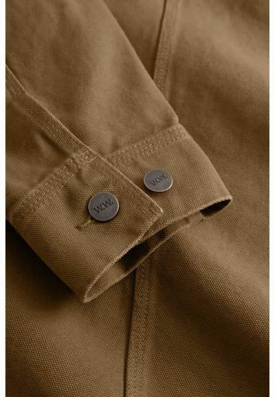 Ruskea puuvillakangas, jossa taitettu hihansuu, jossa on kaksi metallista painiketta, joissa on kaiverrus "WWW." Näkyvissä on ompelu- ja tekstuuridetaljeja.