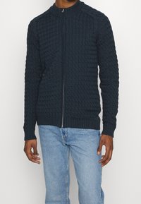 Man som bär en mörk marinblå kabelstickad zip-up tröja med ribbade ärmslut och nederkant i kombination med ljusblå jeans.