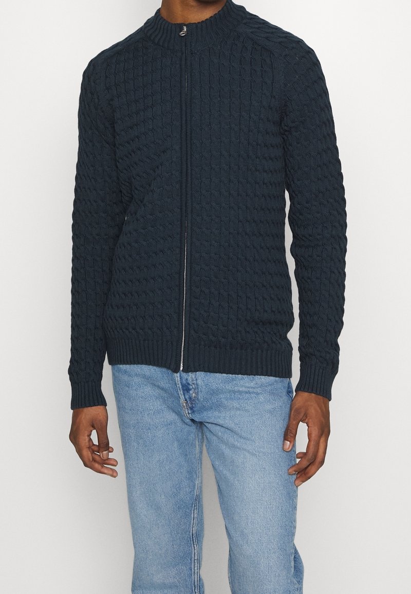 Man som bär en mörk marinblå kabelstickad zip-up tröja med ribbade ärmslut och nederkant i kombination med ljusblå jeans.