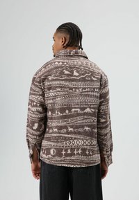 Bruin wollen overhemd met witte geometrische patronen en dierenmotieven, voorzien van een kraag en met knopen gesloten manchetten, weergegeven van achteren.