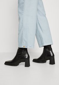 Schwarze Leder-Ankle-Boots mit blockigem Absatz, glatter Textur, spitzer Zehenpartie und seitlichem Reißverschluss, kombiniert mit hellblauen weit geschnittenen Hosen.