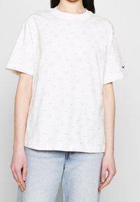 Bílé bavlněné tričko s opakujícím se světlým šedým vzorem kruhů a malých log "Nike", s krátkými rukávy a kulatým výstřihem.