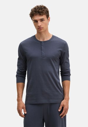Junger Mann mit kurzem lockigem Haar, der ein dunkelblaues Henley-Oberteil mit langen Ärmeln und drei Knöpfen sowie passende Hosen trägt, steht vor hellem Hintergrund.