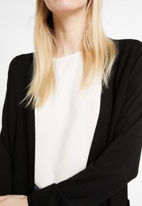 Black cardigan with a soft texture and open front design, worn over a white blouse. Close-up view focuses on the garment details.
