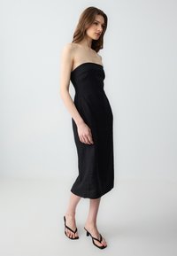Schwarzes, trägerloses Midi-Kleid aus leichtem Material mit körpernaher Silhouette und seitlichen Raffungn. Kombiniert mit schwarzen, hohen Flip-Flops.