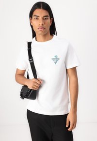 T-shirt blanc à manches courtes avec un logo triangulaire vert et bleu, associé à un pantalon noir et à un petit sac noir avec une chaîne.