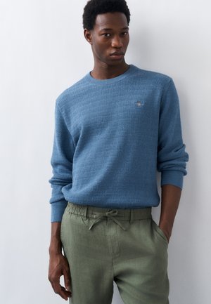 Mężczyzna w niebieskim swetrze o fakturze i oliwkowych spodniach z troczkami, stoi przy białej ścianie, z lewą ręką w kieszeni.