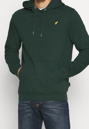 Mann trägt dunkelgrünes Kapuzensweatshirt mit gelbem Vogel-Logo auf der Brust und Hände in der Kängurutasche, kombiniert mit blauen Jeans.