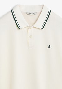Camiseta polo color crema con un cuello a rayas verdes, que presenta un pequeño logo de calavera negra y huesos cruzados en el lado izquierdo del pecho. Tela texturizada.