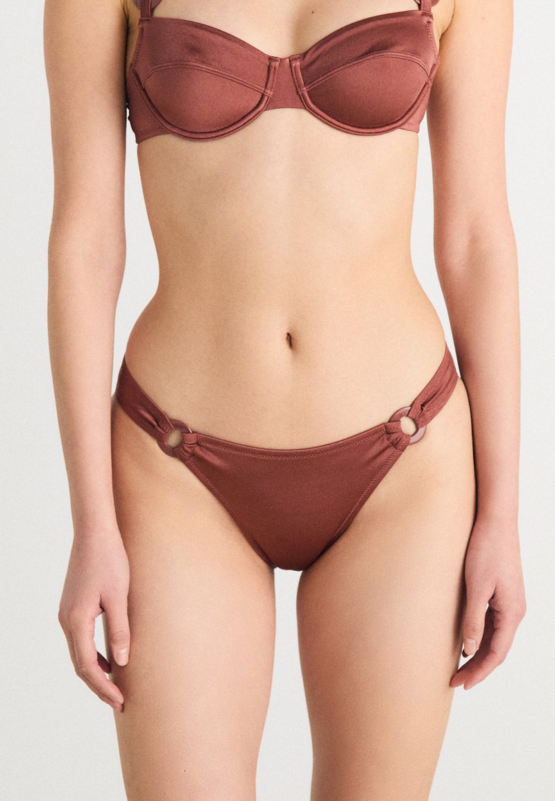 Ensemble bikini marron comprenant un soutien-gorge structuré avec armatures et bretelles réglables, associé à un bas taille basse agrémenté de détails en métal circulaires.