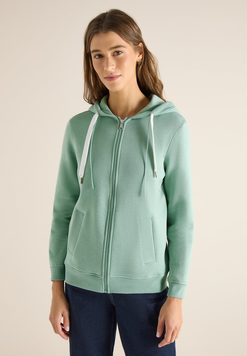 Femme portant un sweat à capuche zippé vert clair avec des cordons blancs, debout contre un fond beige uni, les mains détendues le long du corps.