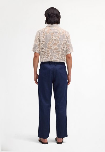 Osoba stojąca tyłem, ubrana w beżową koszulę z krótkim rękawem z koronkowymi wzorami, granatowe spodnie i czarne sandały.