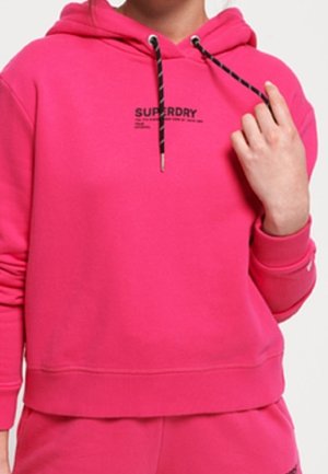 Osoba nosząca jasnoróżową bluzę Superdry z czarno-białymi sznurkami, widoczna od szyi do bioder, trzymająca jeden sznurek.