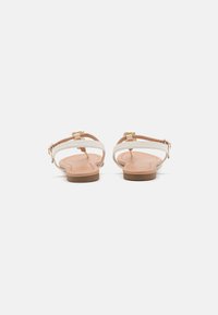 Sandales plates avec une semelle beige clair, dotées de brides blanches et d'accents en métal doré. Design simple avec des straps réglables pour un ajustement parfait.