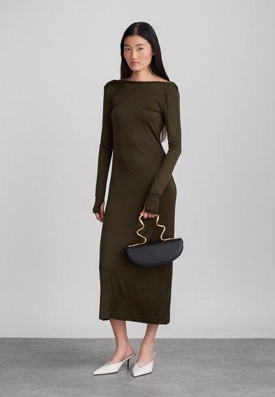 Vestido longo de manga longa verde-oliva com decote barco, combinado com uma bolsa curvada preta com um cabo dourado ondulado e mules brancas de salto.