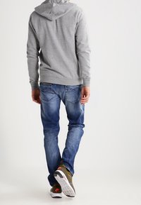 Hombre vestido con sudadera gris, jeans azul y zapatillas Nike verdes oliva caminando hacia atrás sobre un fondo blanco liso.