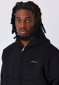 Sudadera con capucha negra con cremallera, con un logo plateado en el pecho, bolsillo tipo canguro y un tejido suave y texturizado.